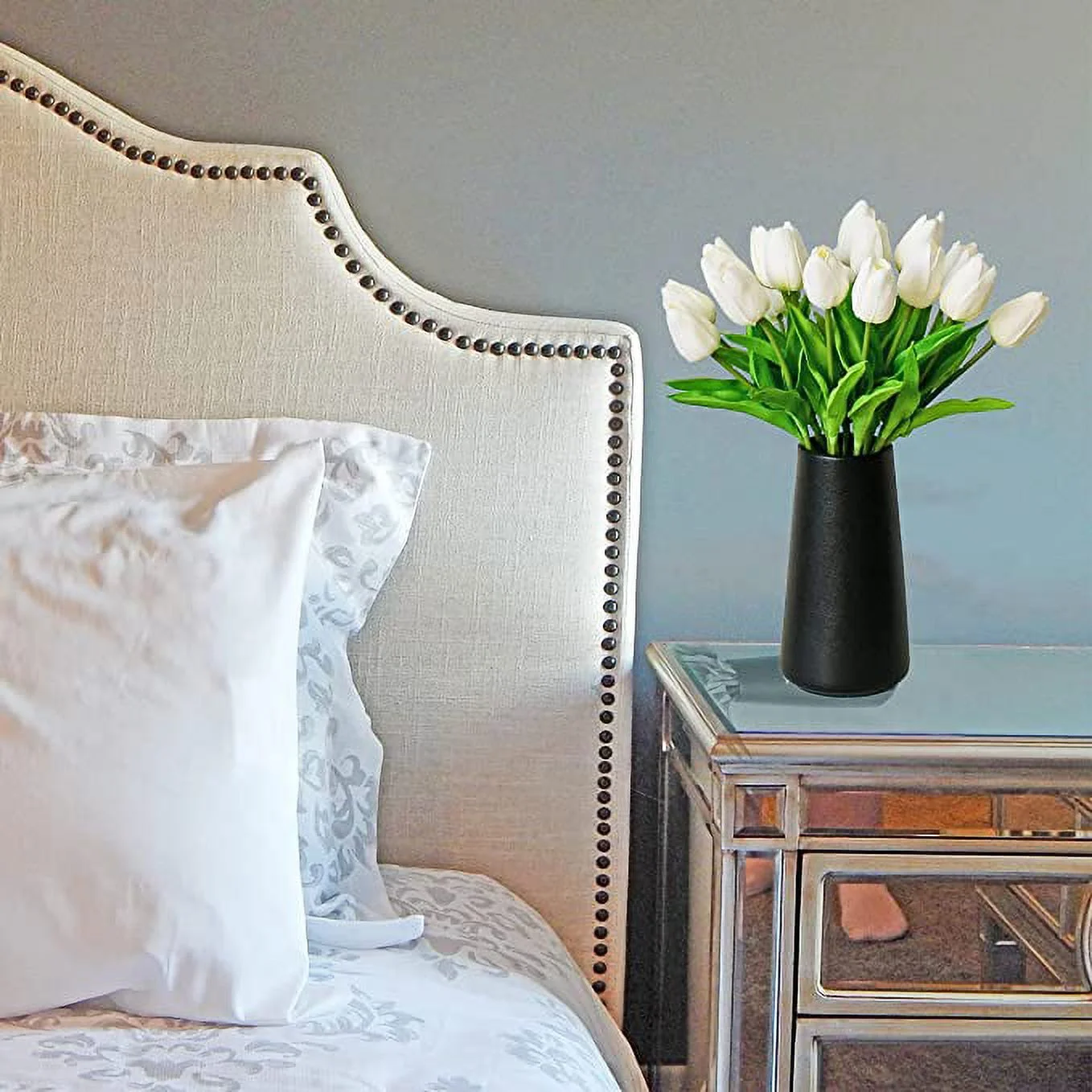White tulips in a black vase on a nightstand next to a bed with white pillows.