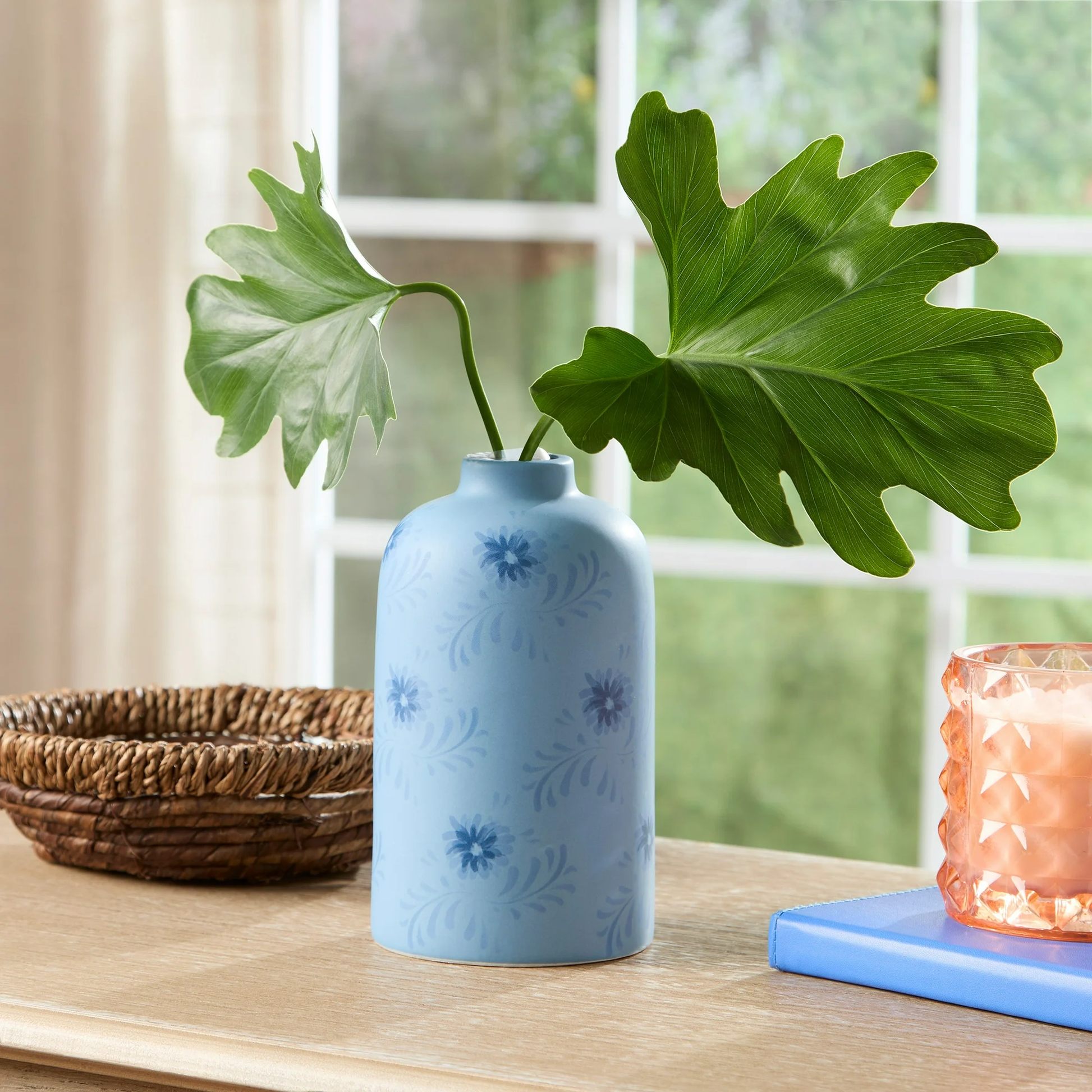 Blue vase with floral patterns on a wooden surface, containing a plant with large green leaves.