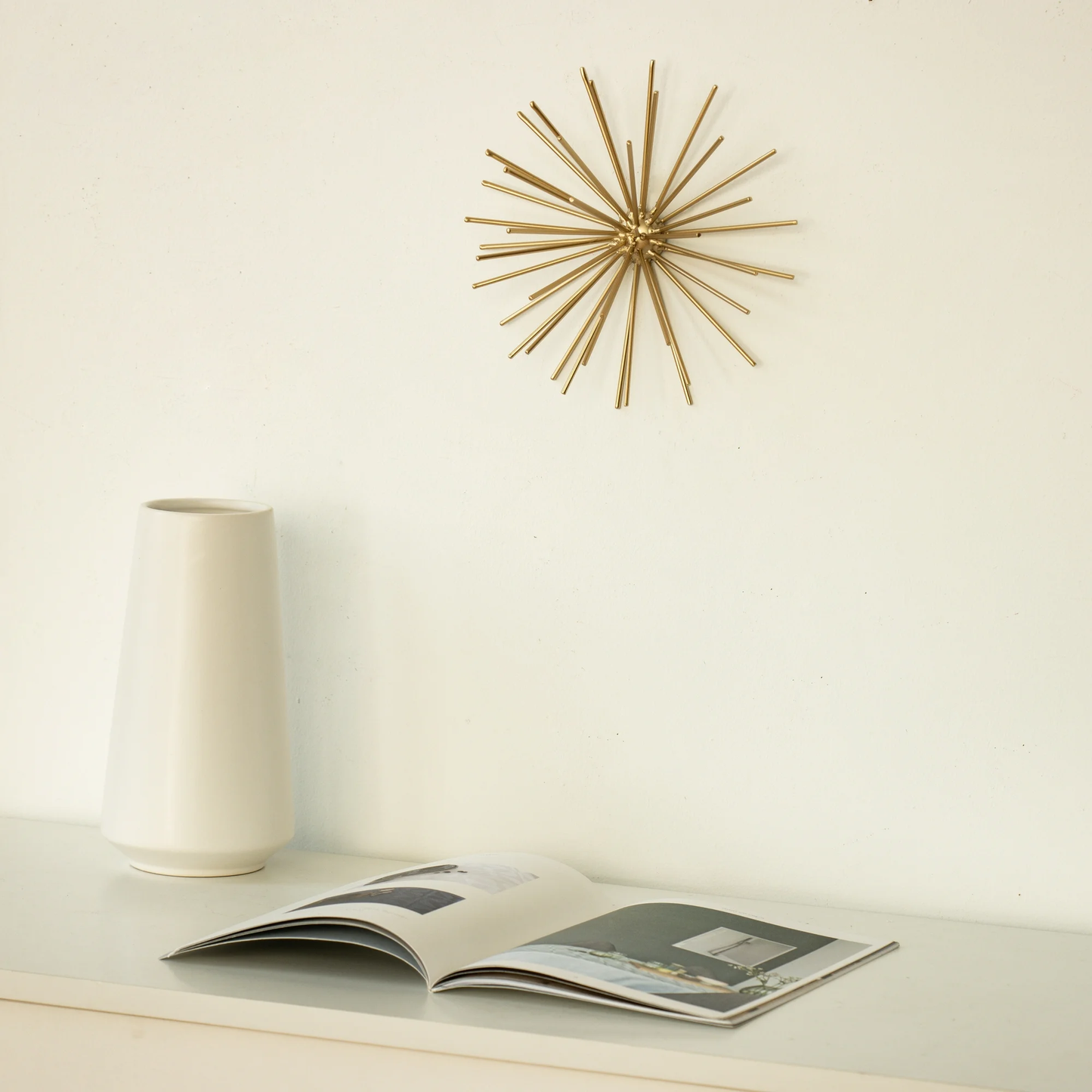 Decorative gold wall clock on a white wall with a vase and open magazine below.