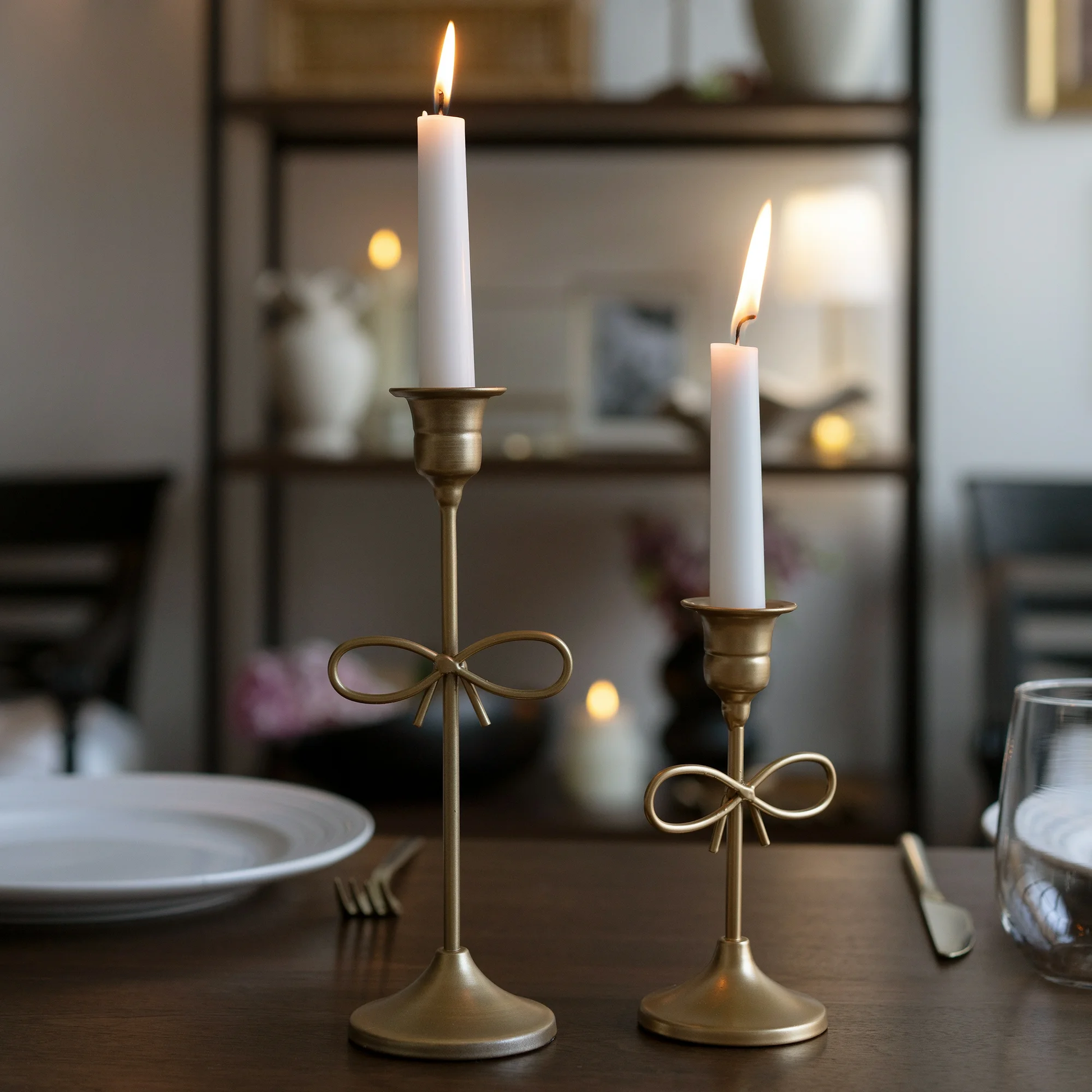 Two gold candle holders with lit candles on a table in a softly blurred indoor setting.